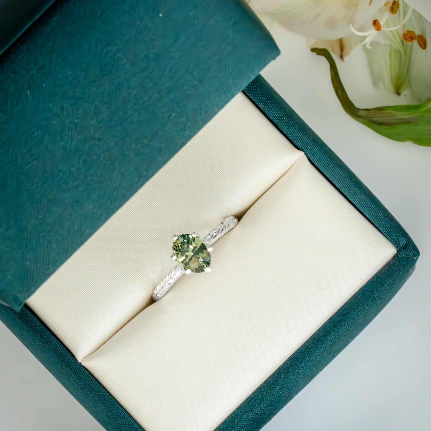 Silver ring with a green gemstone in a teal jewelry box with white flowers in the background